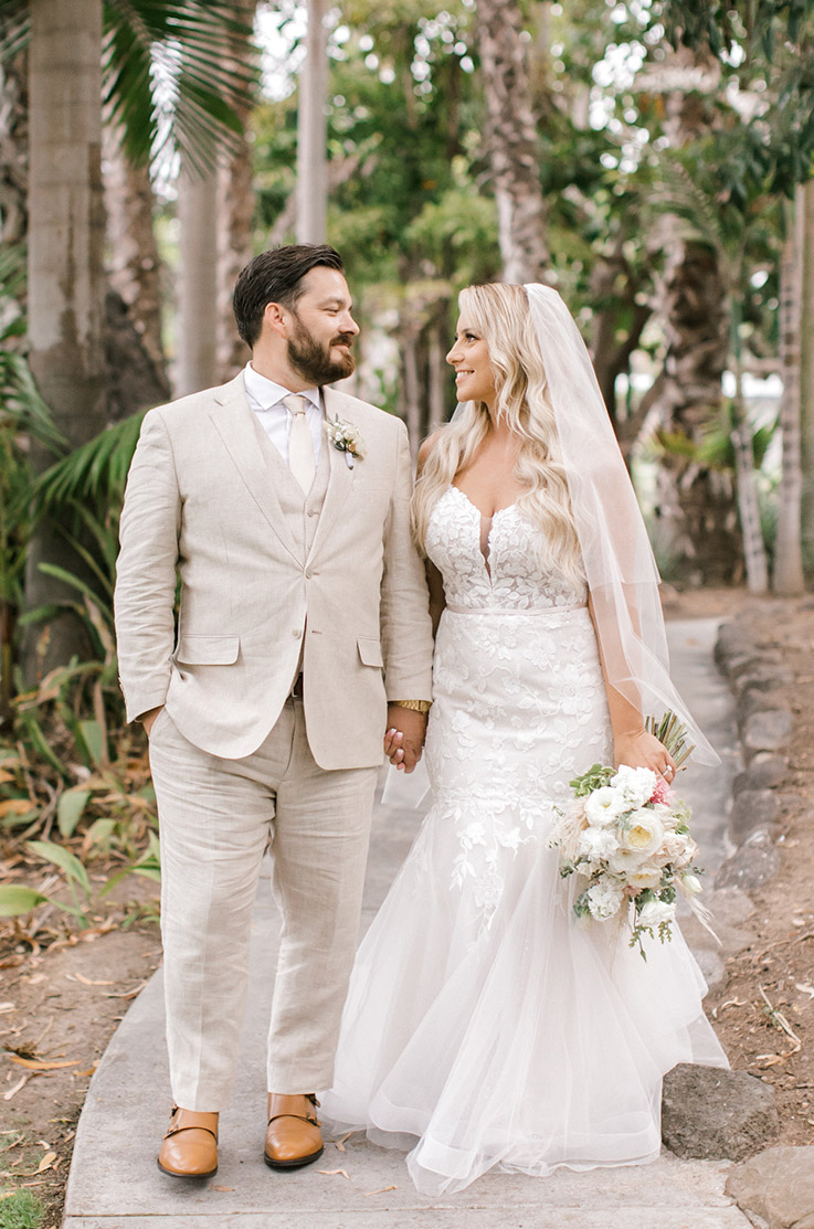 Bride walks in a strapless plunging neck gown with lace details, beside groom in a tan suit on a path lined with palm trees.