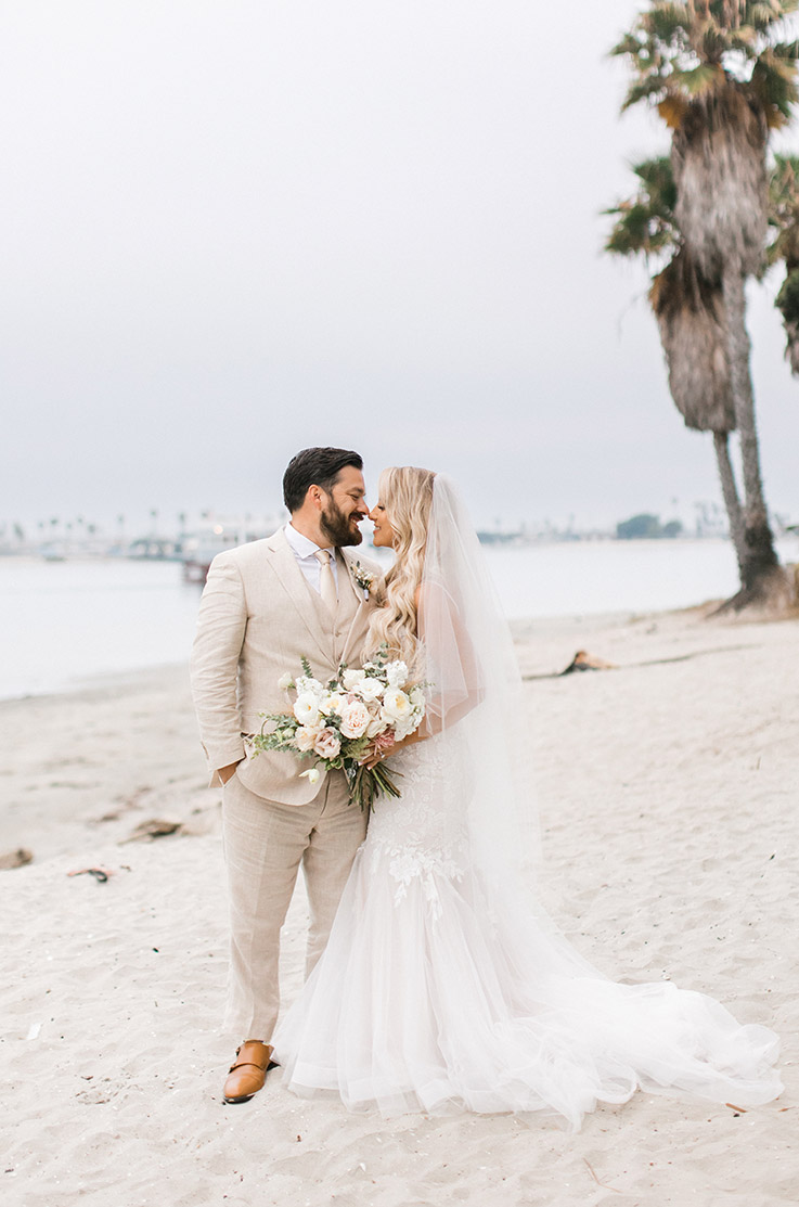 Groom embraces bride in a strapless tulle wedding gown with lace and a veil, holding a blush bouquet on a white sand beach.