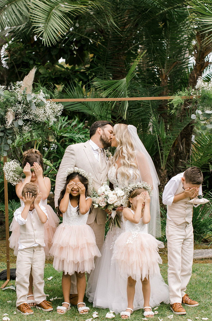 Groom kisses his bride at altar in a strapless lace gown with a tulle skirt and a veil, poses with bridal party by palm trees.