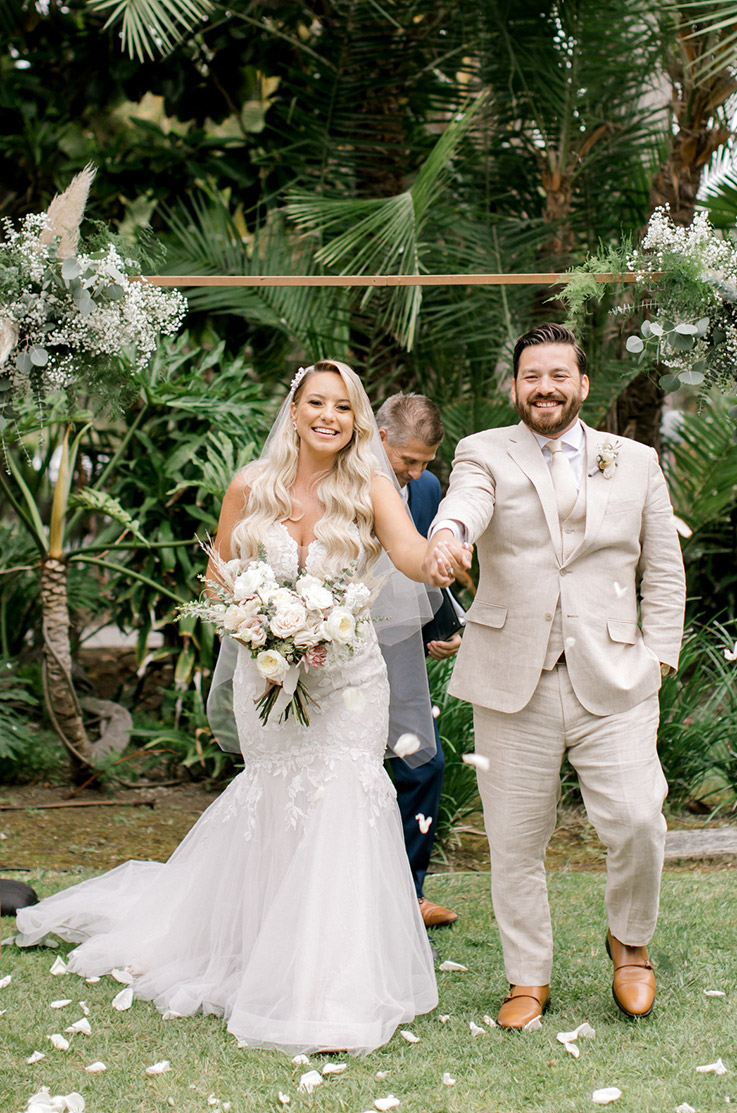 Bride in a plunging neck lace gown with a mermaid tulle skirt, walks alongside groom in the aisle at an outdoor ceremony.