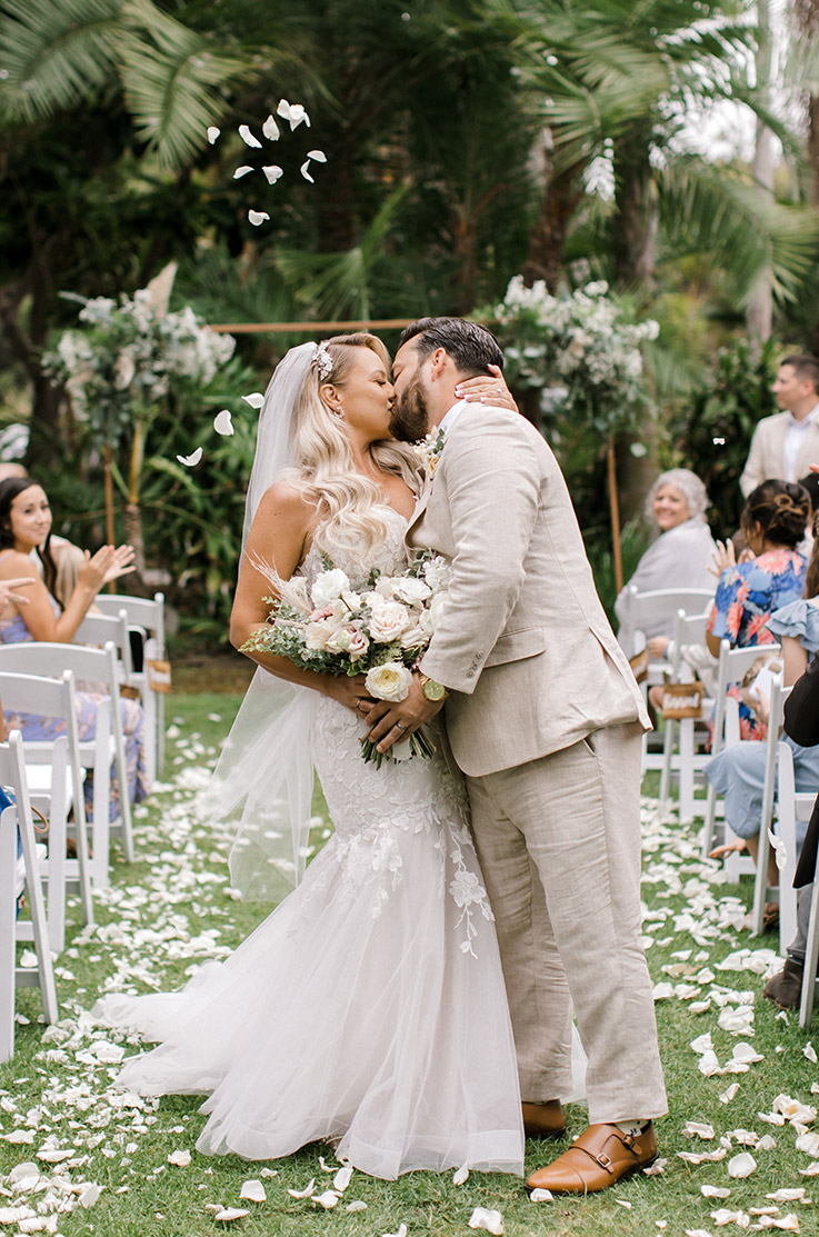 Groom kisses bride in a strapless lace gown, lined in tulle with a sweetheart neck in the aisle in front of tropical plants. 