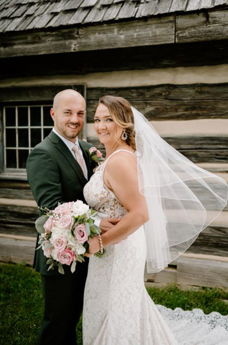 Bride in a sleeveless lace fitted gown with a veil, poses beside her groom outside a log cabin, holding a blush bouquet.