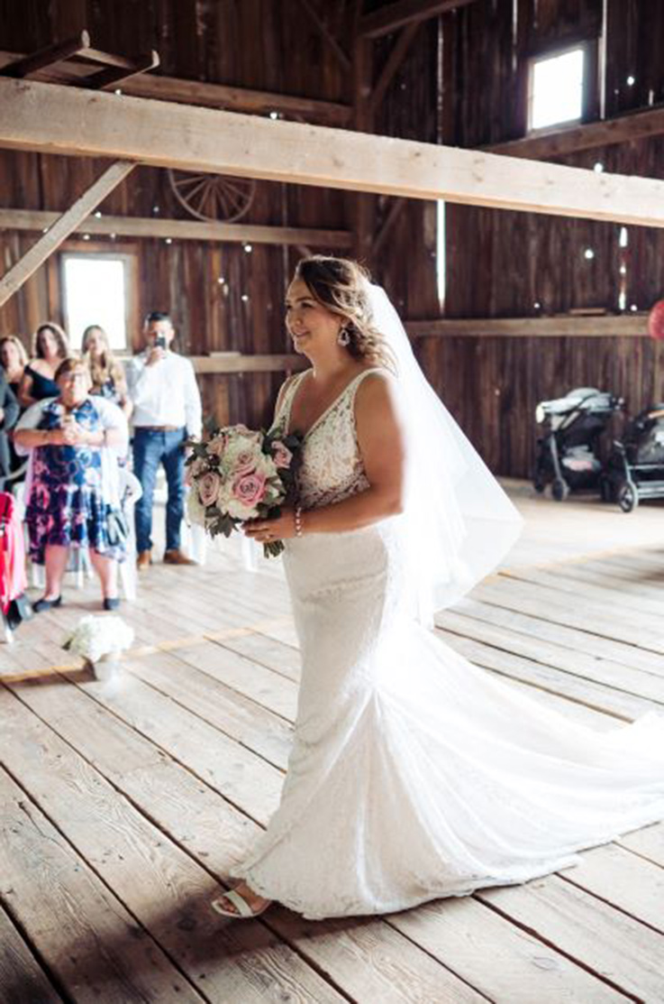 Bride in a lace fit and flare wedding dress with a plunging neck, walks down the aisle in a wooden barn, holding a bouquet.