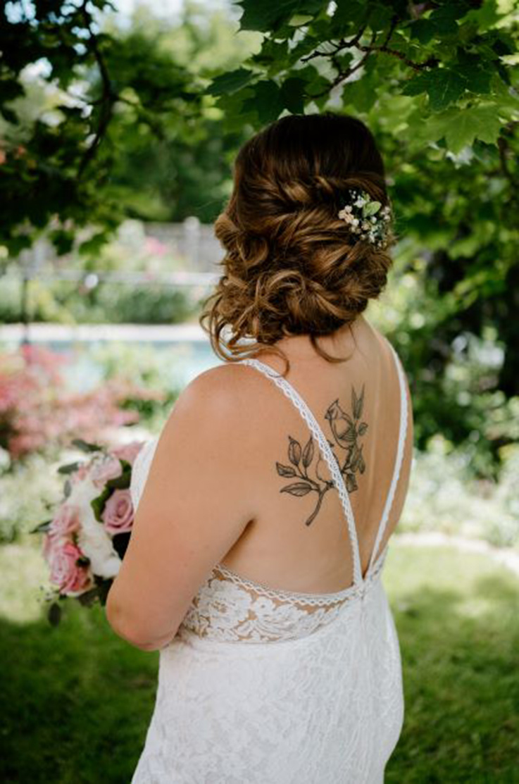 Back of bride in a lace gown with a low back and lace trim detail, is holding a pink and white bouquet underneath trees.