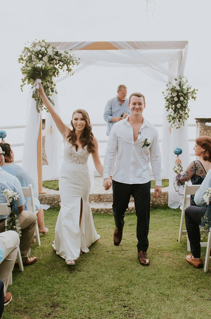Bride in a lace gown with a fit and flare crepe skirt and a V-neck, holds a bouquet up and walks next to groom in the aisle. 