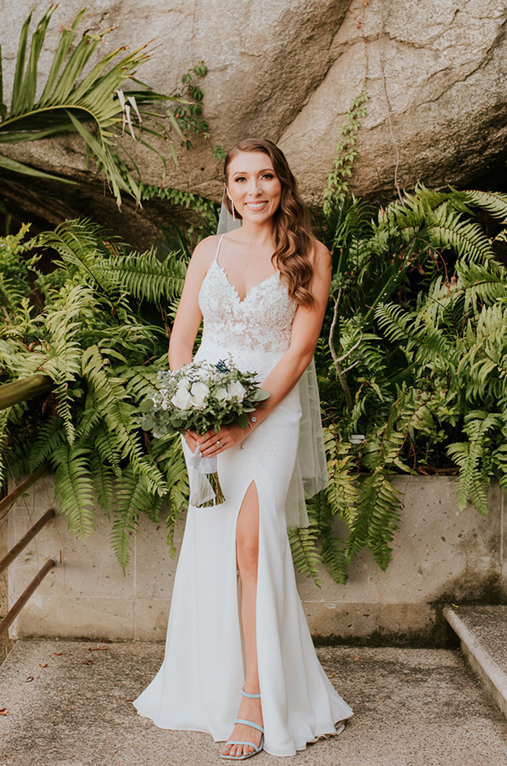 Bride in a lace V-neck wedding dress with a crepe skirt, a skirt slit and veil, holds a bouquet on steps with tropical plants.