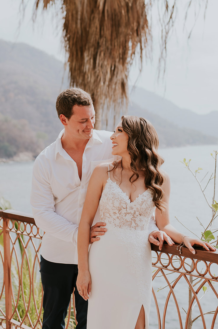 Bride in a spaghetti strap lace gown with a crepe fit and flare skirt and skirt slit, looks up at her groom on an oceanside terrace.