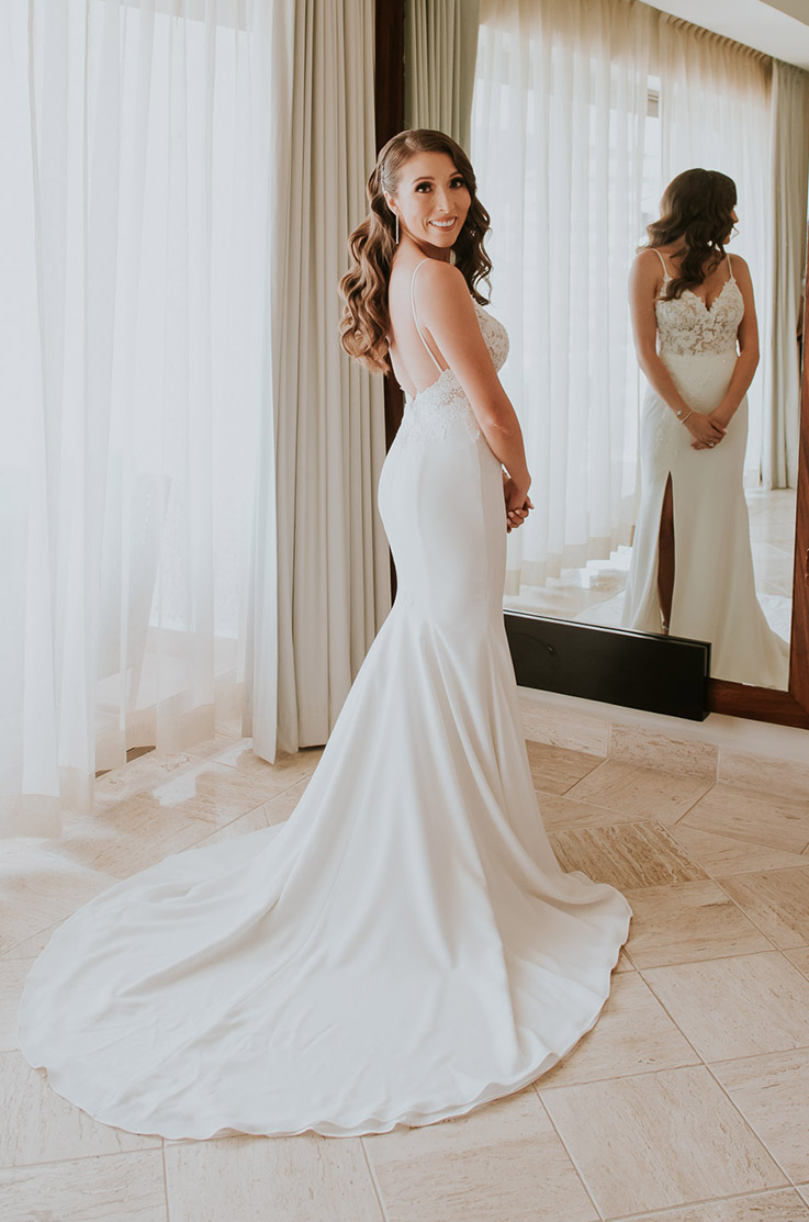 Bride looks over her shoulder in a low back lace gown with a fit and flare crepe skirt, standing at a mirror in bright room.