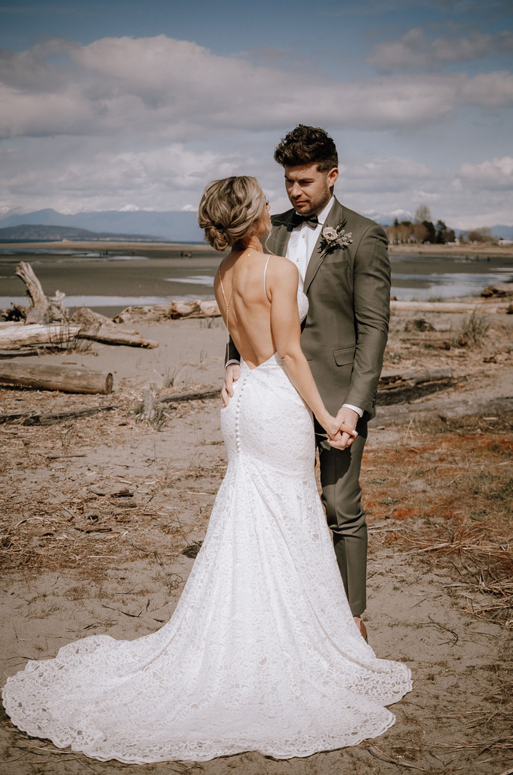 Groom poses with bride in an open back lace wedding dress with fit and flare skirt and buttons, on a beach with mountain views.