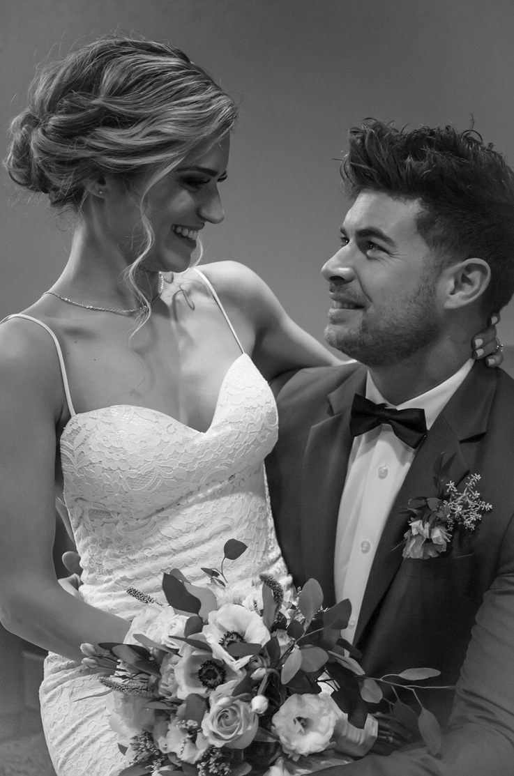 Bride in a lace sweetheart neck gown, faces groom and is holding a bouquet on his lap in black and white portrait.