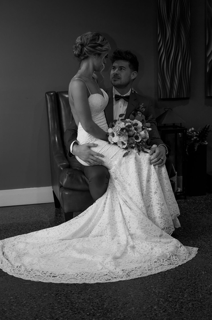 Bride sits in an all lace gown with a sweetheart neck and holds a bouquet, on a leather chair with groom in an indoor setting.