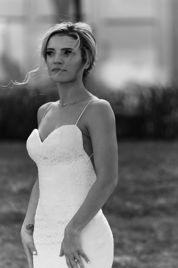 Close-up of bride looking ahead in a lace sweetheart neck gown with thin straps, is standing outdoors and in black and white.