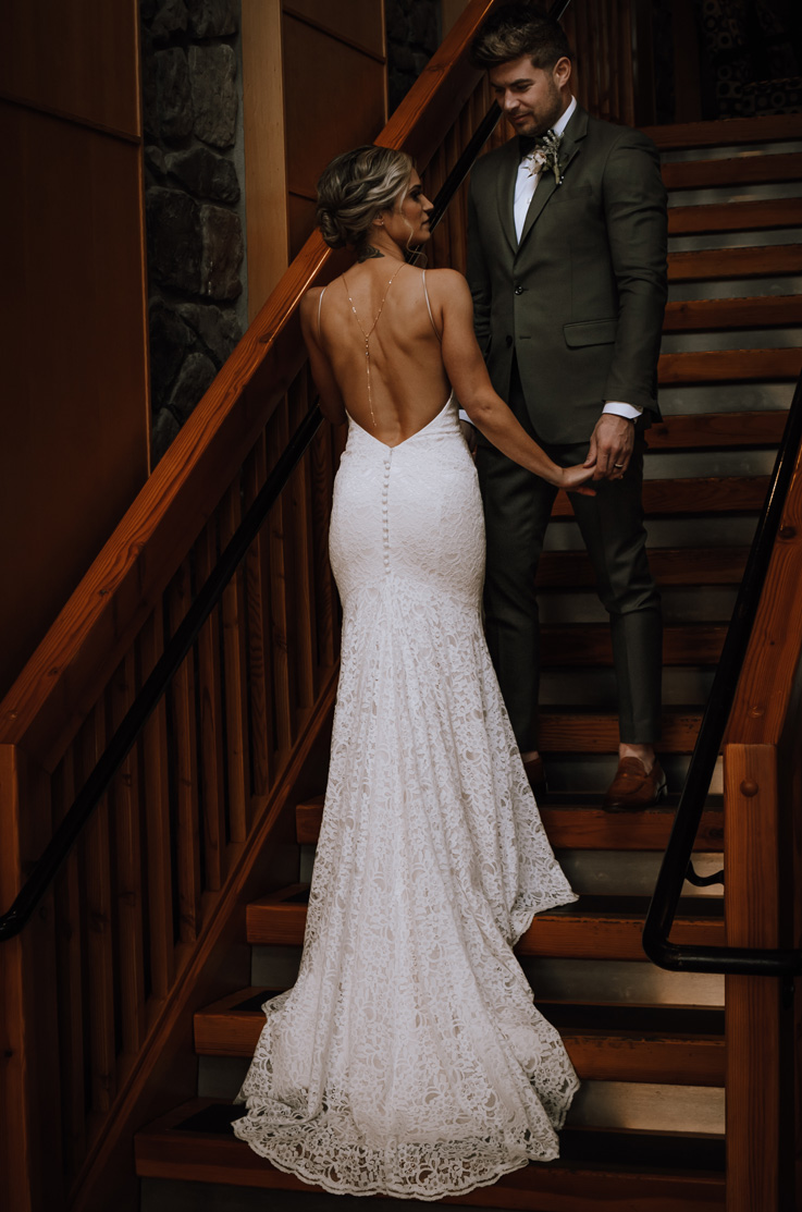 Bride walks up stairs in a backless lace gown with buttons and a fit and flare skirt, in front of groom in dark lighting.