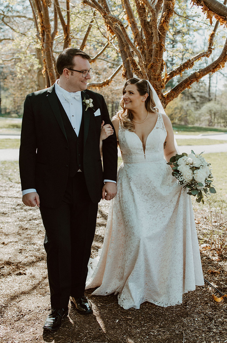 Bride in a sleeveless lace gown with double bands across the waist, holds a white bouquet and walks beside groom outdoors.