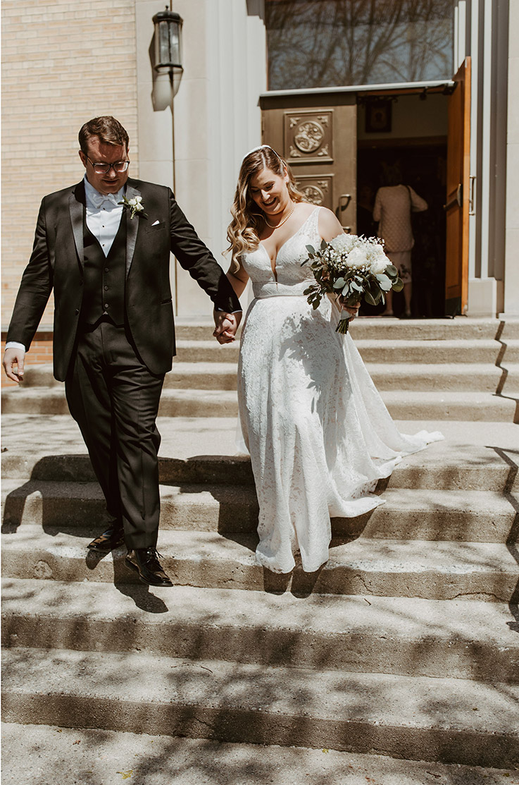 Bride in a lace A-line wedding dress with double beaded bands and a plunging neck, walks next to groom on church steps.