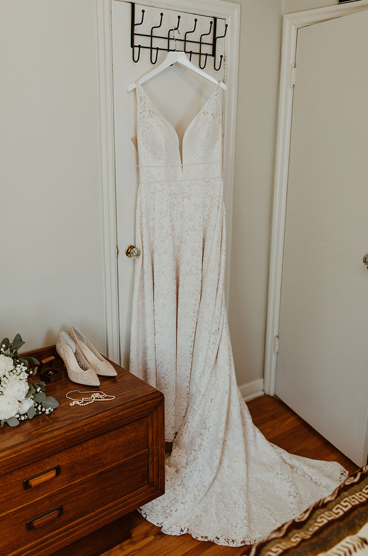A sleeveless lace A-line gown with a plunging neck bodice and double thin bands, hangs on the back of a doorway indoors. 