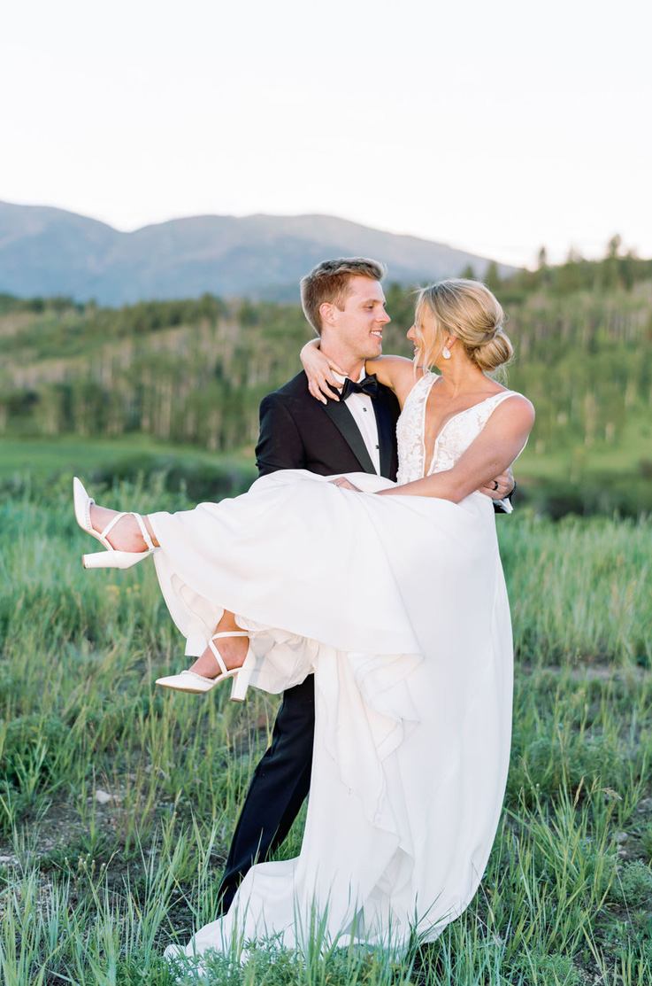 Groom carries his bride in a lace bodice wedding dress with a crepe skirt and plunging neck, standing in a vast green meadow. 