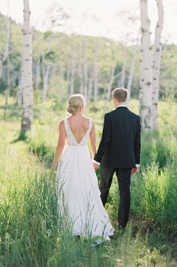 Bride in an open back lace gown with a full crepe skirt, walks hand in hand with the groom through a sunny forest clearing.