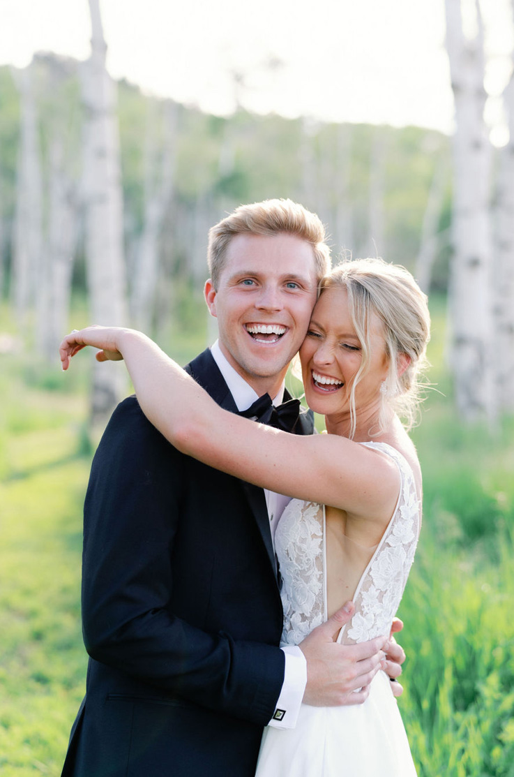Groom laughs with his bride in an embrace, wearing a sleeveless lace bodice gown with side cutouts, in a sunny forest area.
