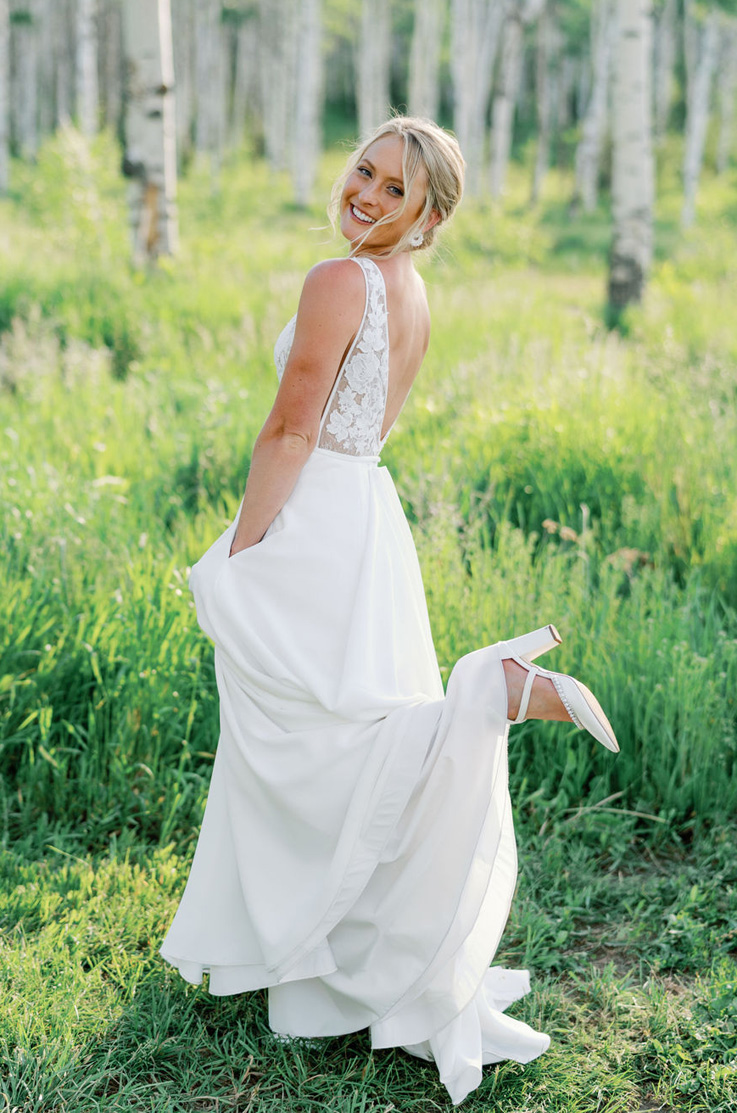 Bride poses in a sunny meadow in a low back and sleeveless lace gown with a crepe skirt, in front of tall grass and trees. 