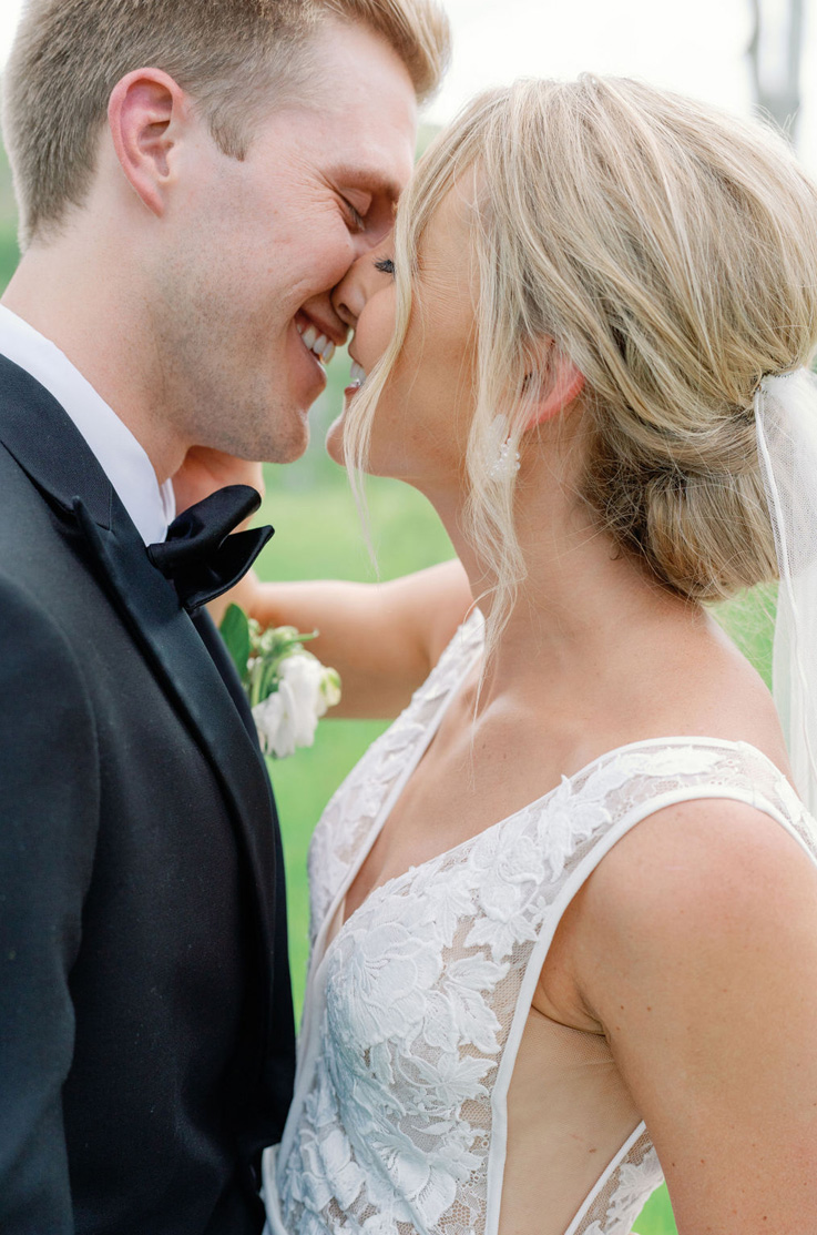Close-up of groom sharing a kiss with his bride in a plunging neck lace gown with side inserts and a veil, smiling outdoors. 