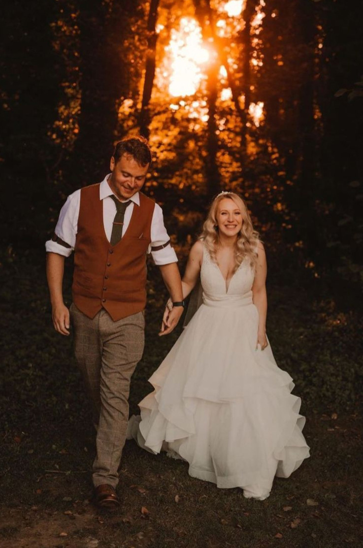 Bride in a V-neck lace wedding dress with a layered tulle skirt and thin bands, walks hand in hand with groom in a forest.