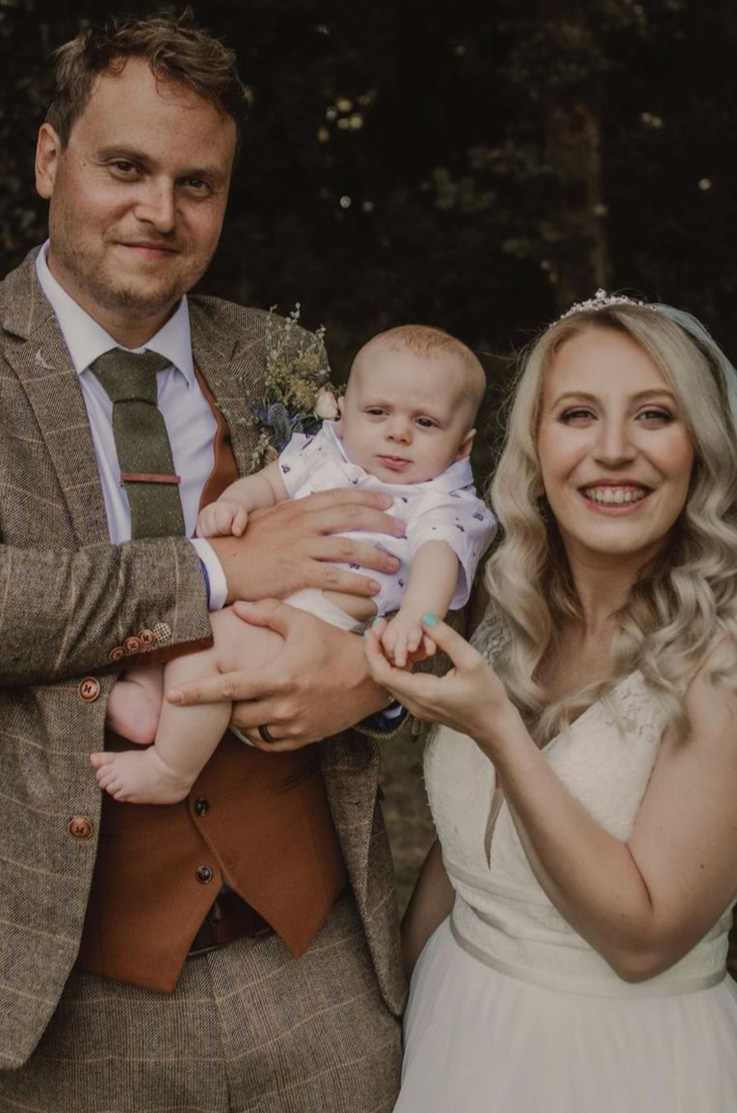 Bride in a V-neck lace gown with thin bands and a flowing A-line skirt, poses beside groom holding their baby in dark lighting.