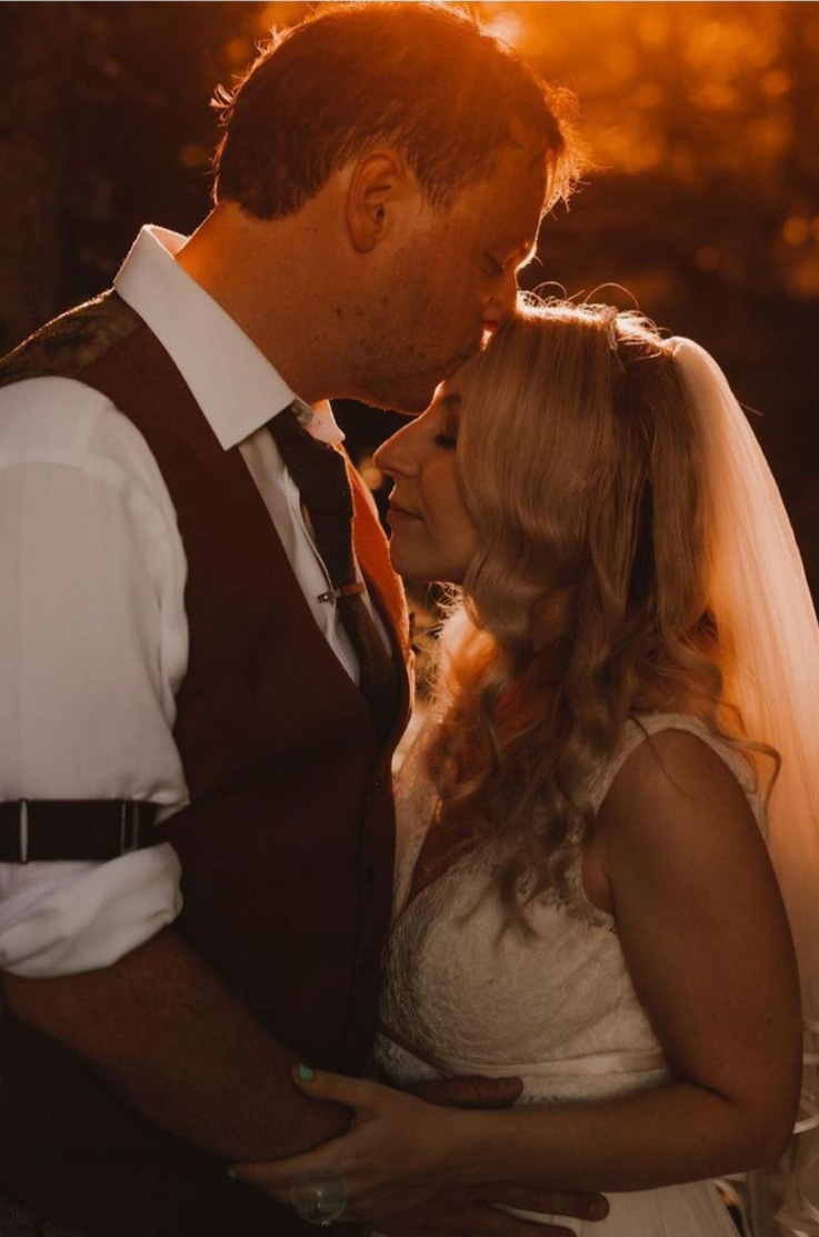 Close-up of groom embracing his bride in a lace bodice gown with a V-neckline, thin bands and a veil, outdoors at golden hour.