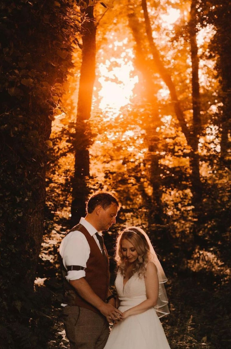 Bride in a lace bodice gown with a V-neckline, a tulle skirt and thin waistbands, poses next to groom at golden hour in a forest.