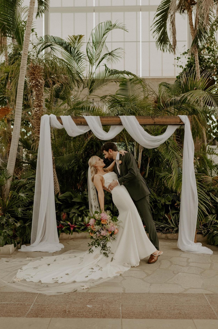 Groom dips his bride at the altar in a crepe scoop neck gown with a buttoned fit and flare skirt and a long veil near palm trees.