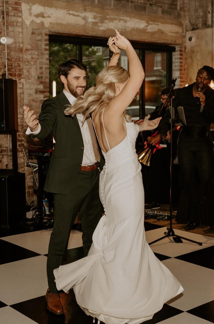 Groom twirls bride in a fit and flare crepe wedding dress with a low back, pleating and buttons, on a checkered dance floor.