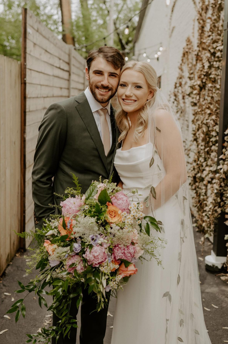 Bride in a pleated crepe bodice gown with a scoop neckline and embroidered veil, is holding a bouquet next to groom outdoors. 