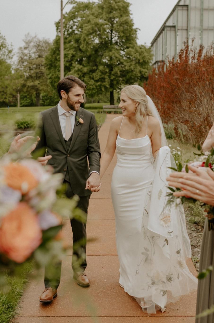Bride in a crepe fit and flare gown with thin straps and an embroided veil, walks hand in hand with groom, facing one other. 