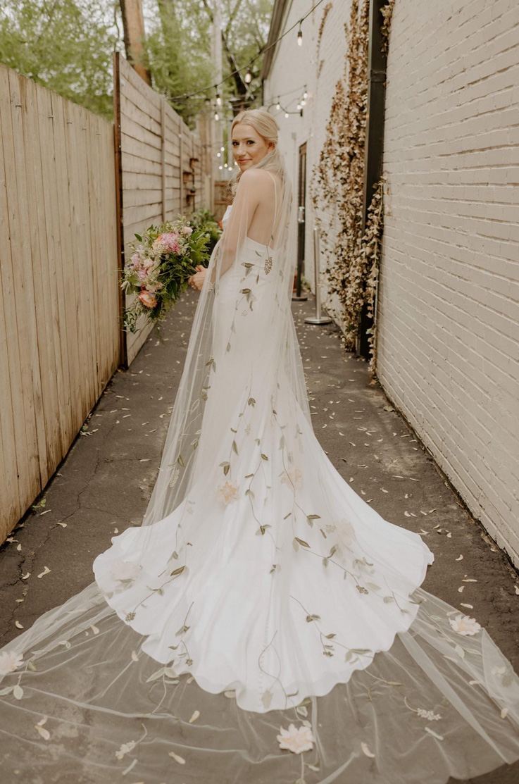  Bride poses in a low back crepe gown with fit and flare skirt and a floral embroidered veil, holds a bouquet outside the venue.