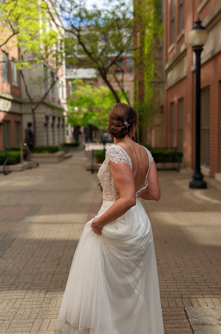 Bride holds her train in a city courtyard, wearing a sheer embroidered wedding dress with a tulle skirt and plunging neckline. 