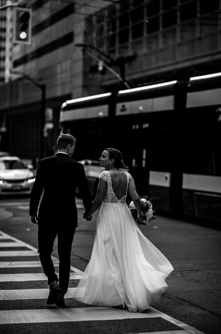 Bride in a low open back tulle gown with sequin appliqueing and cap sleeves, walks hand in hand with groom on a city street.