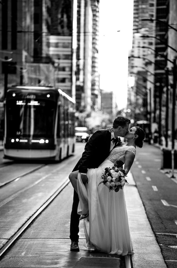 Groom dips bride and picks up her leg in an embroidered tulle gown with a mesh tulle skirt with a slit, standing on a city street. 