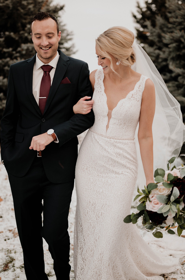A bride holding a bouquet, wearing a plunging neckline lace wedding dress and veil, holds the arm of a groom in a suit as they walk outside and smile, with trees in the background.