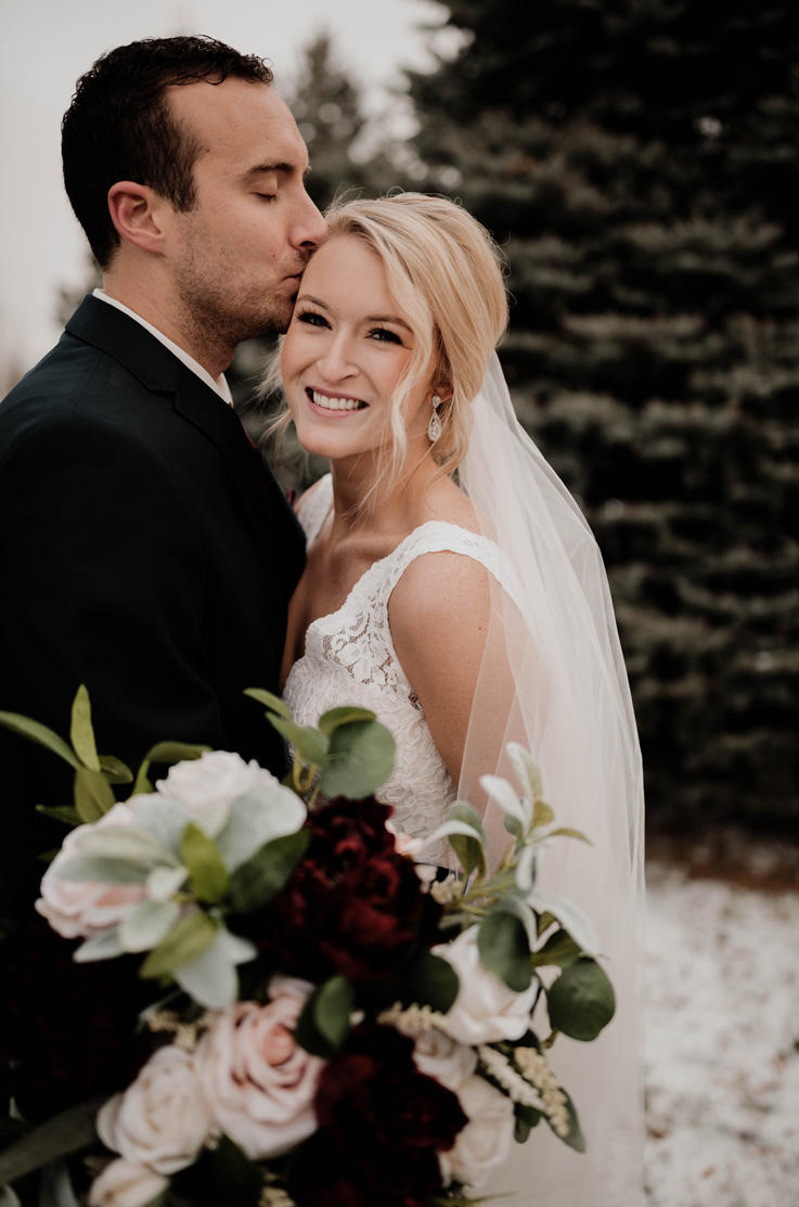 A groom in a suit kisses a smiling bride in a sleeveless lace wedding dress and veil on the head, they both stand outside with trees in the background.