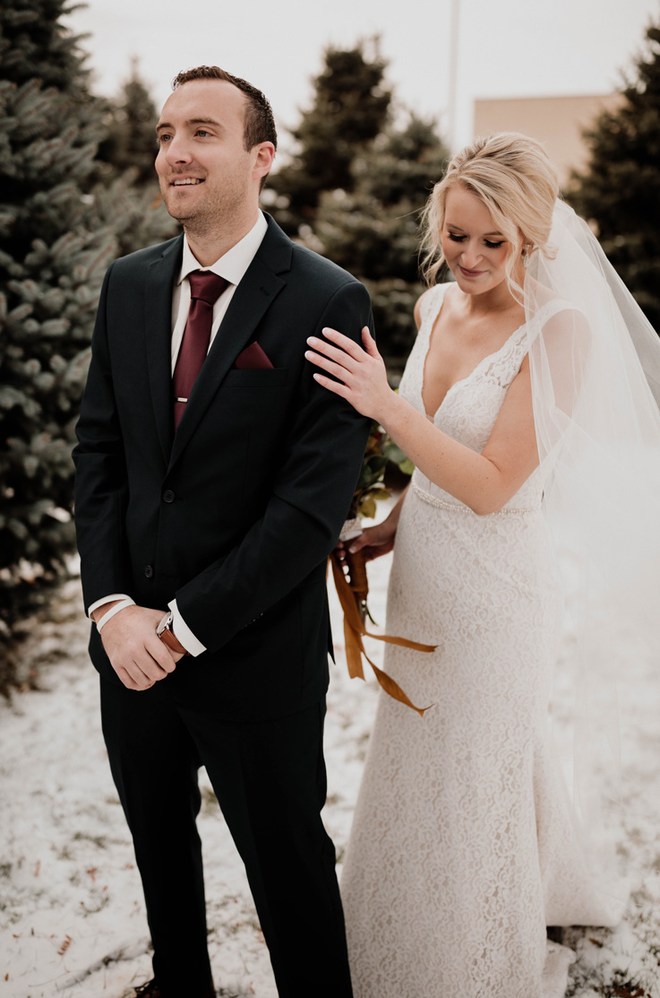 A bride in a sleeveless lace wedding dress and veil touches the arm of a groom in a suit as she approaches him from behind outside, with trees in the background.