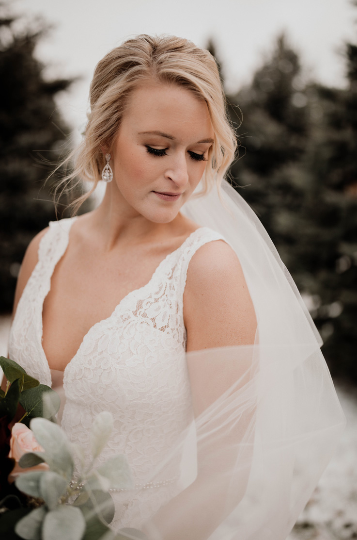 A bride in a sleeveless lace wedding dress and veil looks down and to the side while standing outside and holding a bouquet.