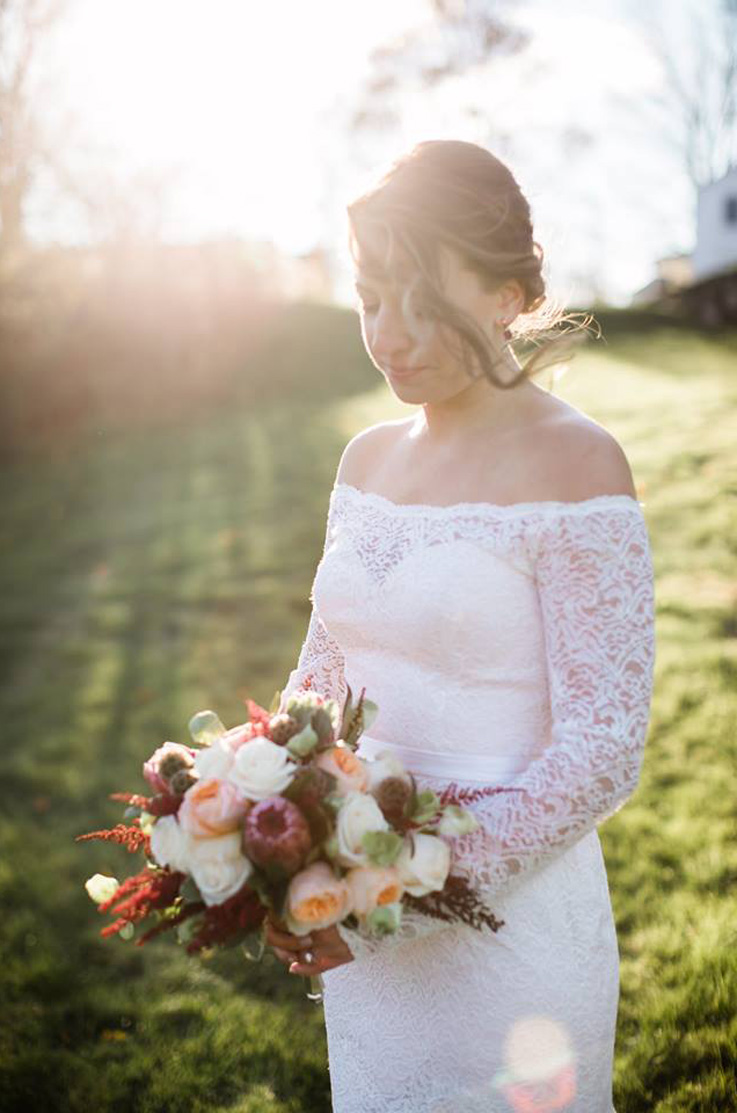 A bride in a long sleeve lace wedding dress holds a bouquet as she stands outside in a field of grass, with the sun shining brightly behind her.