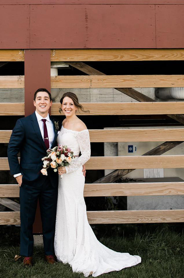 A bride in a long sleeve lace wedding dress holding a bouquet, and a groom in a suit, both smile and stand outside in front of a building with wood slats behind them.