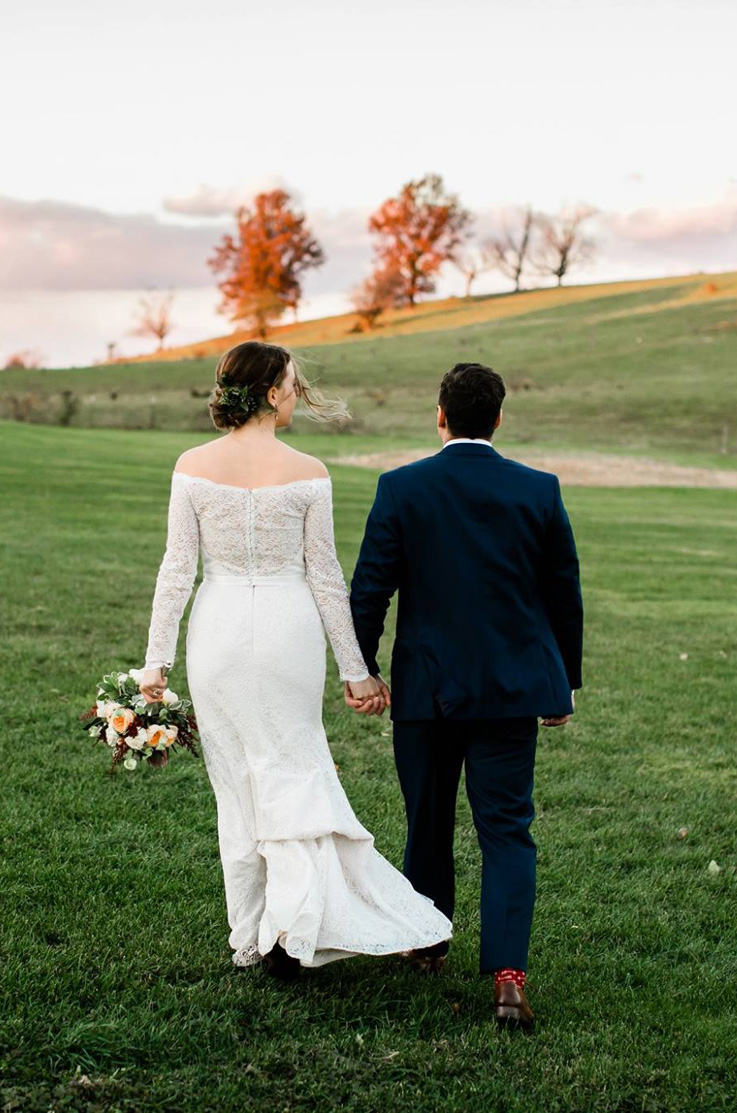 A bride in a long sleeve lace wedding dress and a groom in a suit hold hands and walk outdoors in a grassy field towards a grassy hill with a few trees on it.