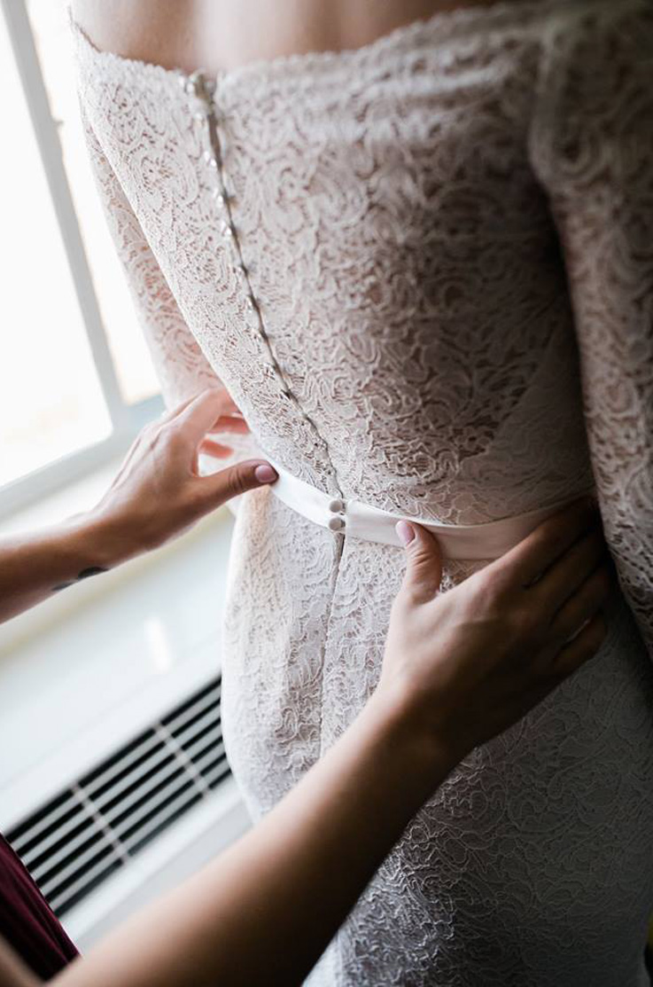 Someone's hands are helping to straighten out the back of a bride's fabric belt, the bride is standing inside and wearing a lace wedding dress with crystal buttons down the back.