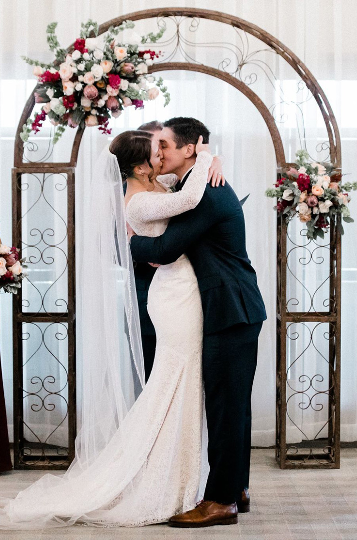 A bride in a long sleeve lace wedding dress and a groom in a suit kiss as they stand in front of a floral decorated wedding arch and sheer curtains.