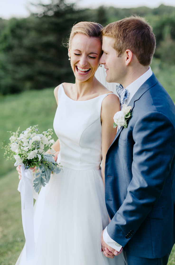 A bride in a sleeveless ball gown wedding dress, holding a bouquet, smiles and laughs as a groom in a suit whispers in her ear, while they stand outside with trees in the background.