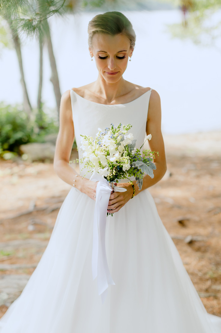 A bride in a sleeveless ball gown wedding dress holds a bouquet and stands outdoors, with a lake and trees behind her.