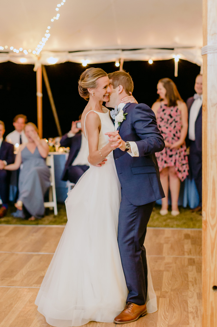 A bride in a sleeveless ball gown wedding dress and a groom in a suit dance together in an outdoor dancefloor tent, with wedding guests watching in the background.