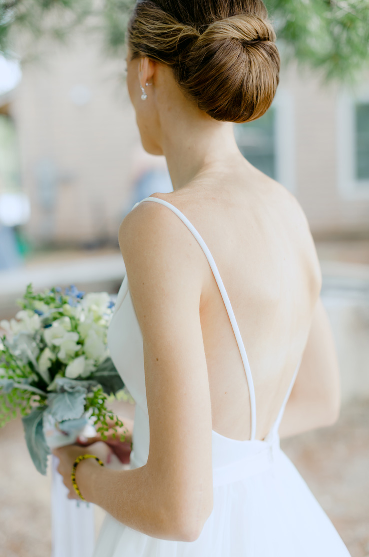 A bride in a wedding dress with spaghetti straps in the back holds a bouquet and stands outside, with a house in the background.